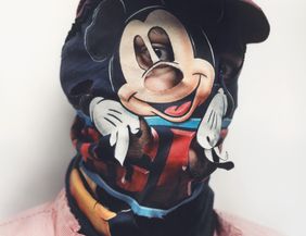 Eine Porträtaufnahme bis zu den Schultern. Das Gesicht ist durch eine weinrote Mütze und vor allem durch eine Stoffmaskierung verdeckt, die die Trickfilmfigur Mickey Mouse zeigt.